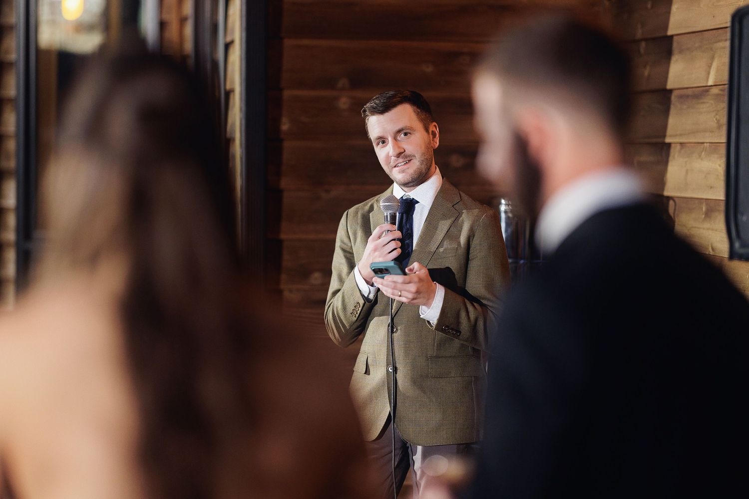 Man in olive suit holding microphone giving speech at elegant wooden interior wedding reception.