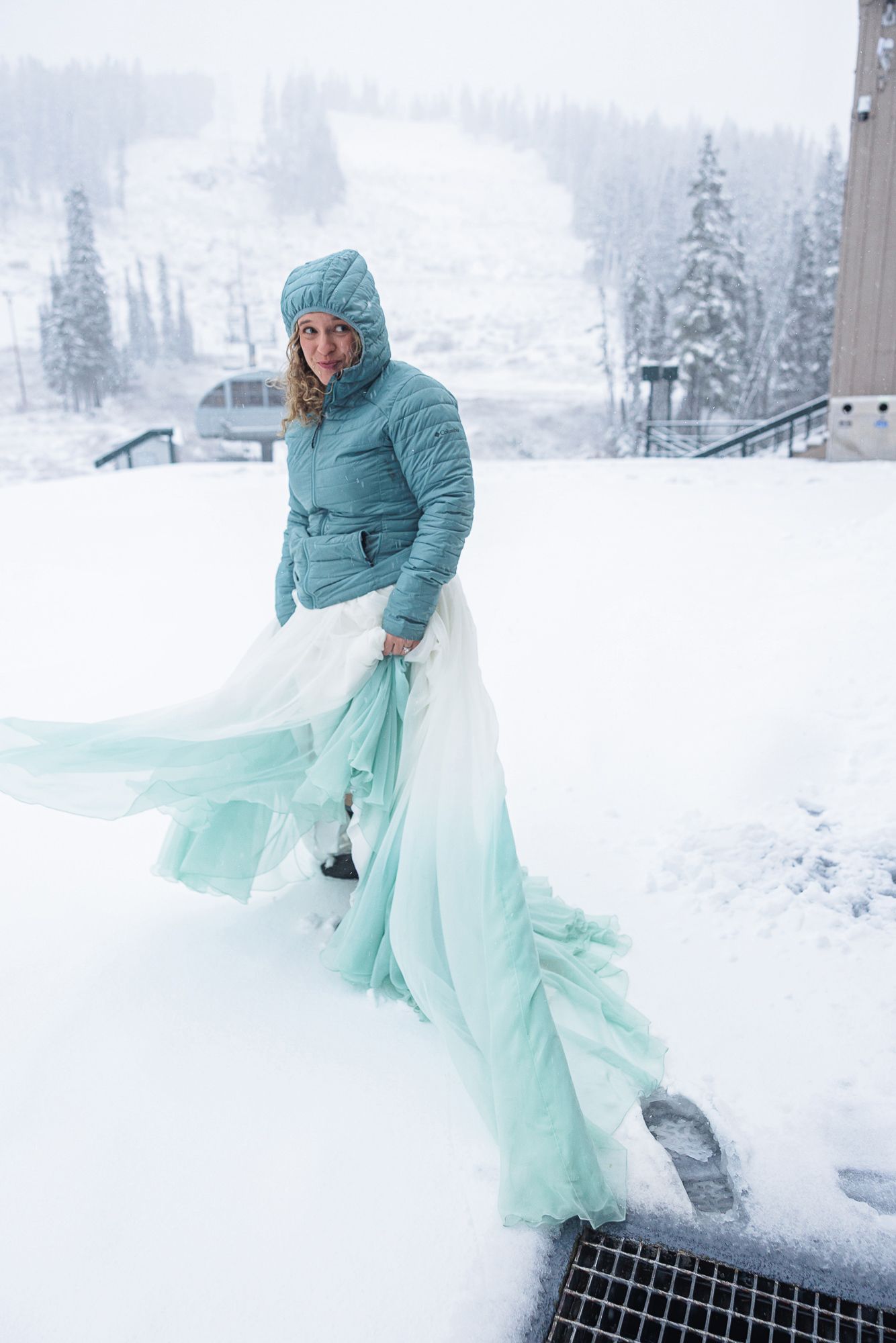 Person in turquoise winter coat and flowy mint dress twirls in snowy winter landscape with evergreen trees.