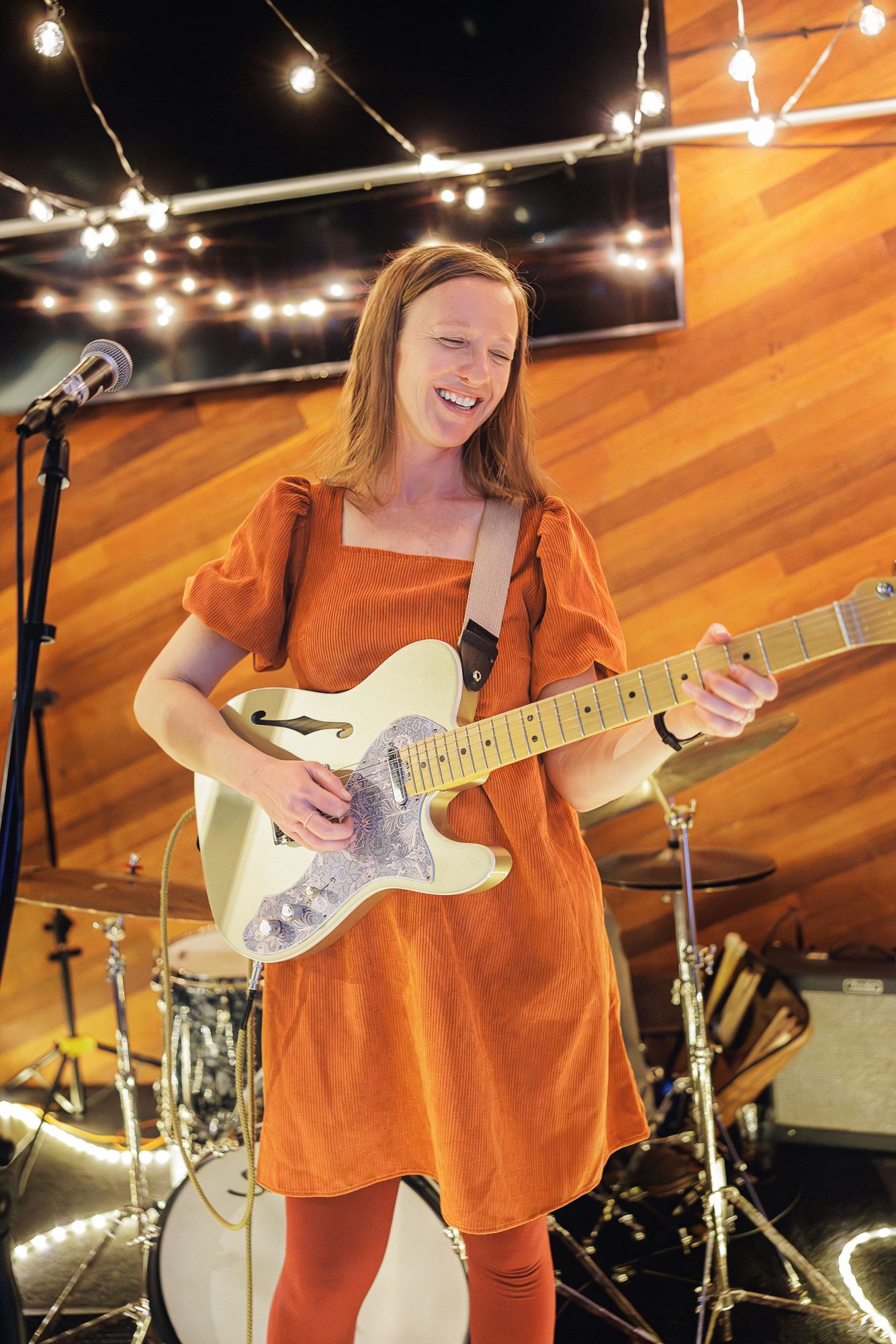 Musician in orange outfit performs on stage with electric guitar under string lights at night.