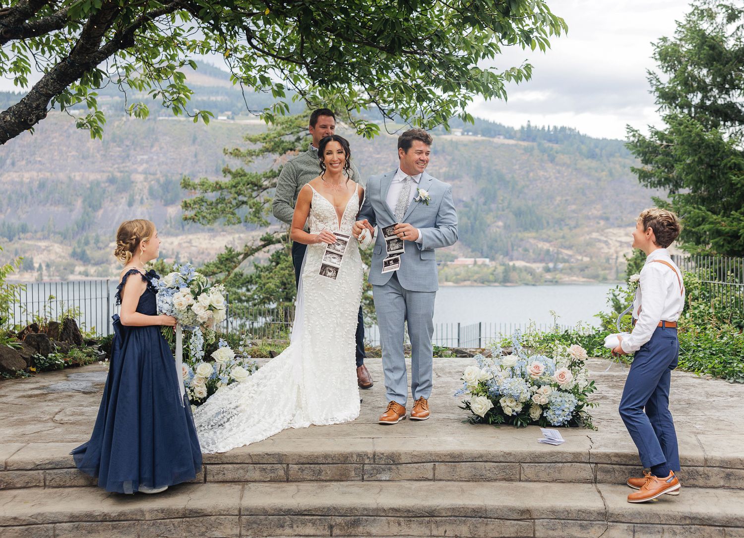 A wedding ceremony takes place on a scenic overlook with mountains and a lake in the background.