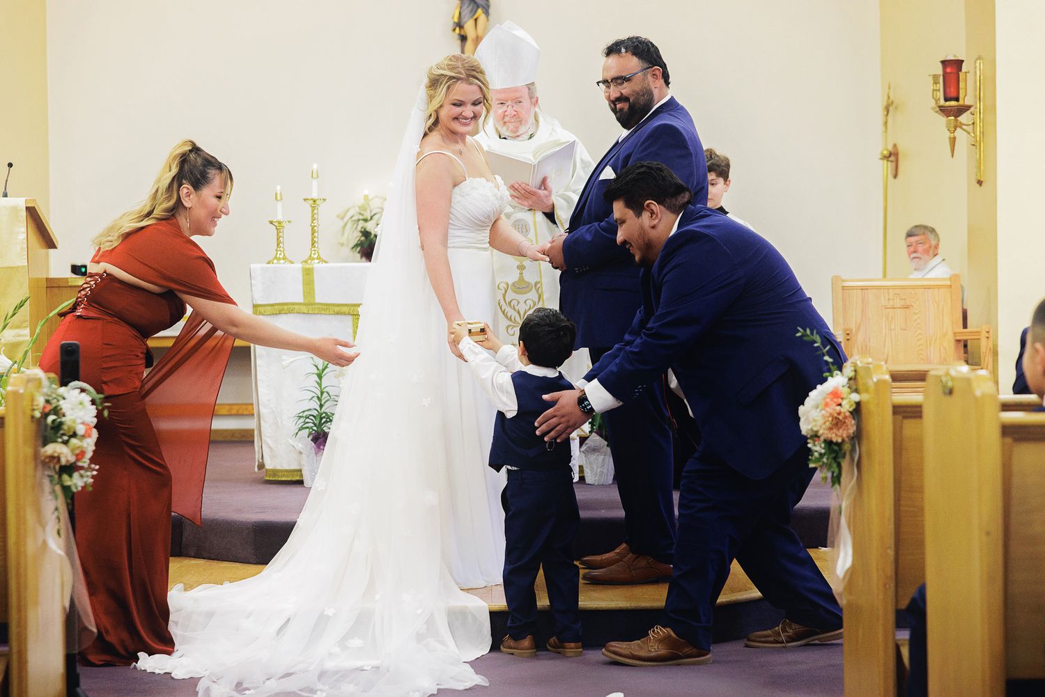 Bride and groom exchange vows at church altar with officiant, wedding party, and ring bearer during ceremony.