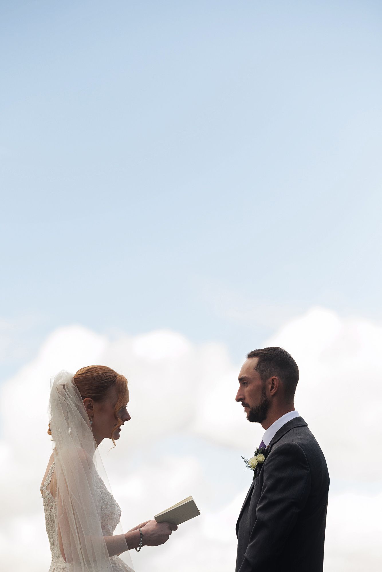 Bride reading wedding vows from a book to groom outdoors against a bright blue sky with white clouds.