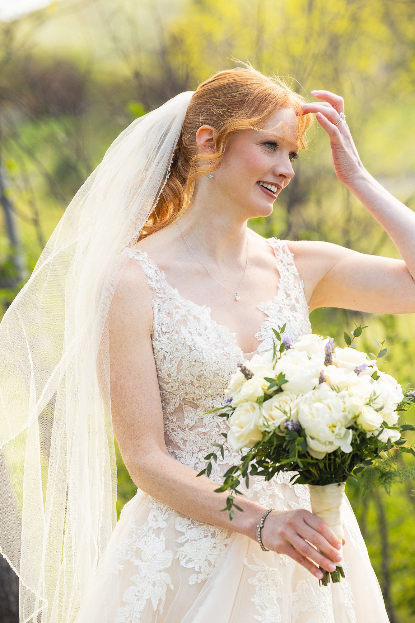 Smiling redhead bride in lace gown holding white bouquet with veil outdoors.