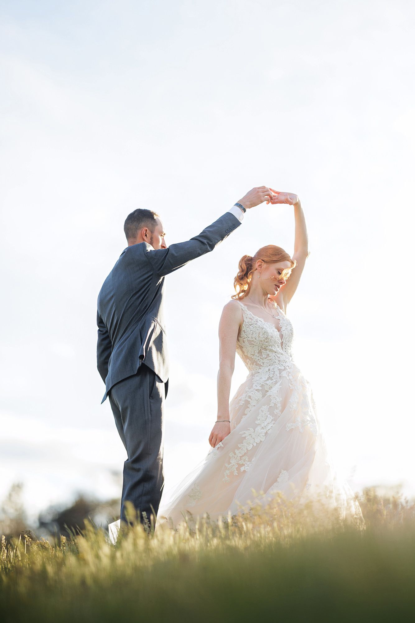 Groom spinning bride in floral wedding gown outdoors in golden hour light on a grassy hill.