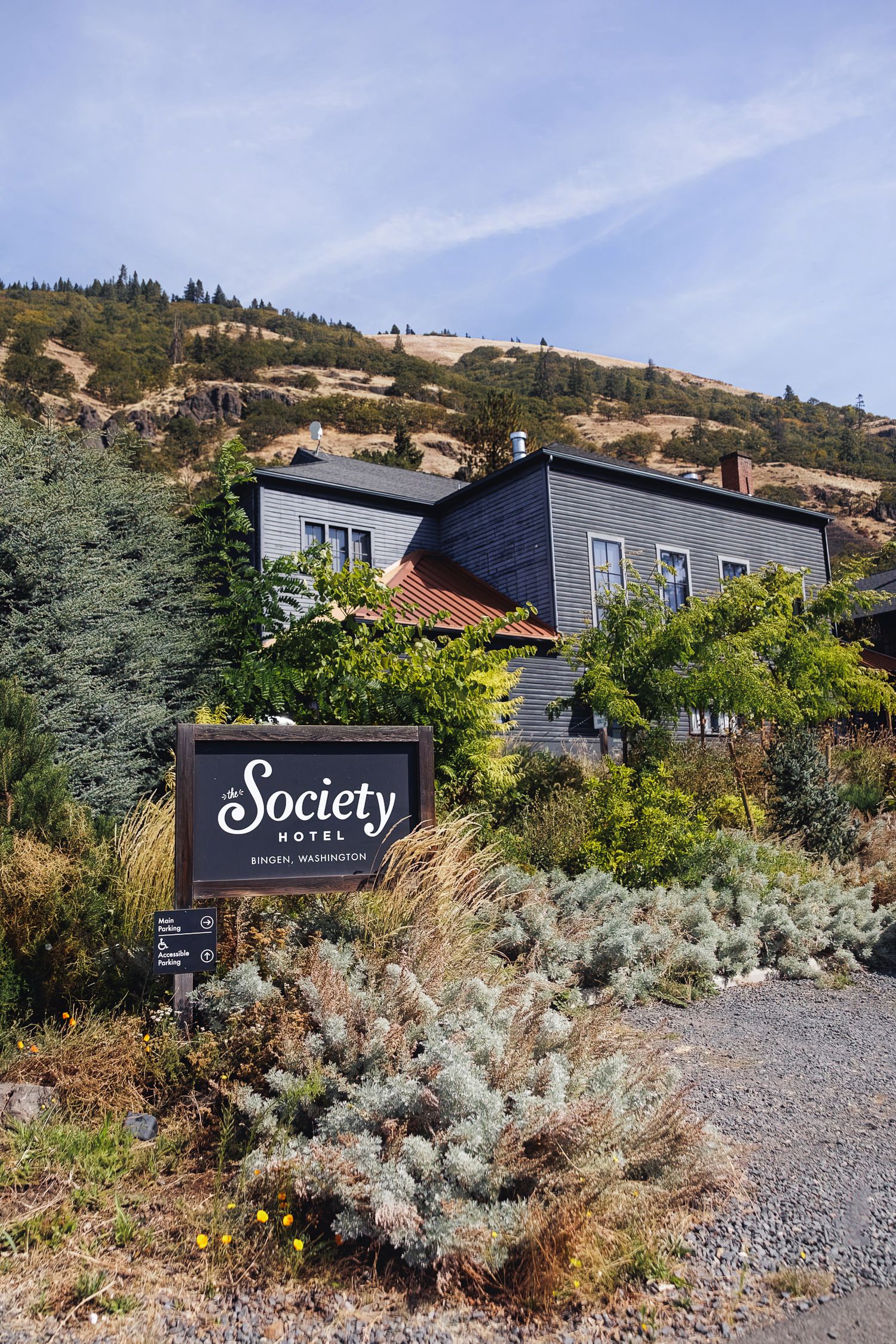 Society Hotel sign in Bingen, Washington, surrounded by native plants with a dark gray building and hillside backdrop.