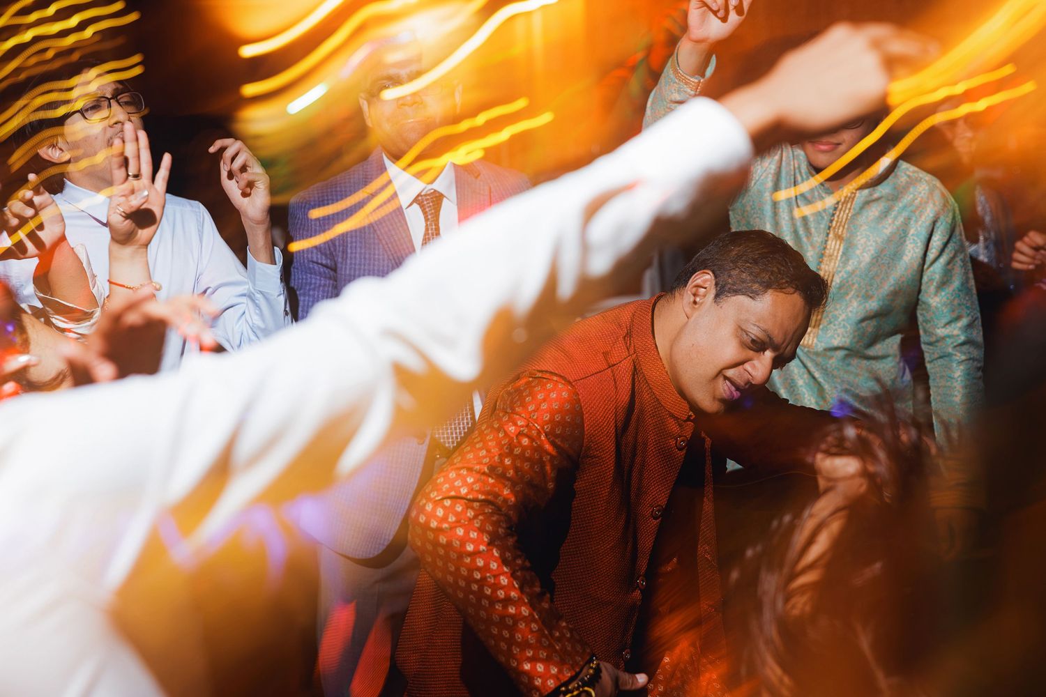 Guests dancing energetically at a vibrant Indian wedding reception with warm orange lighting.