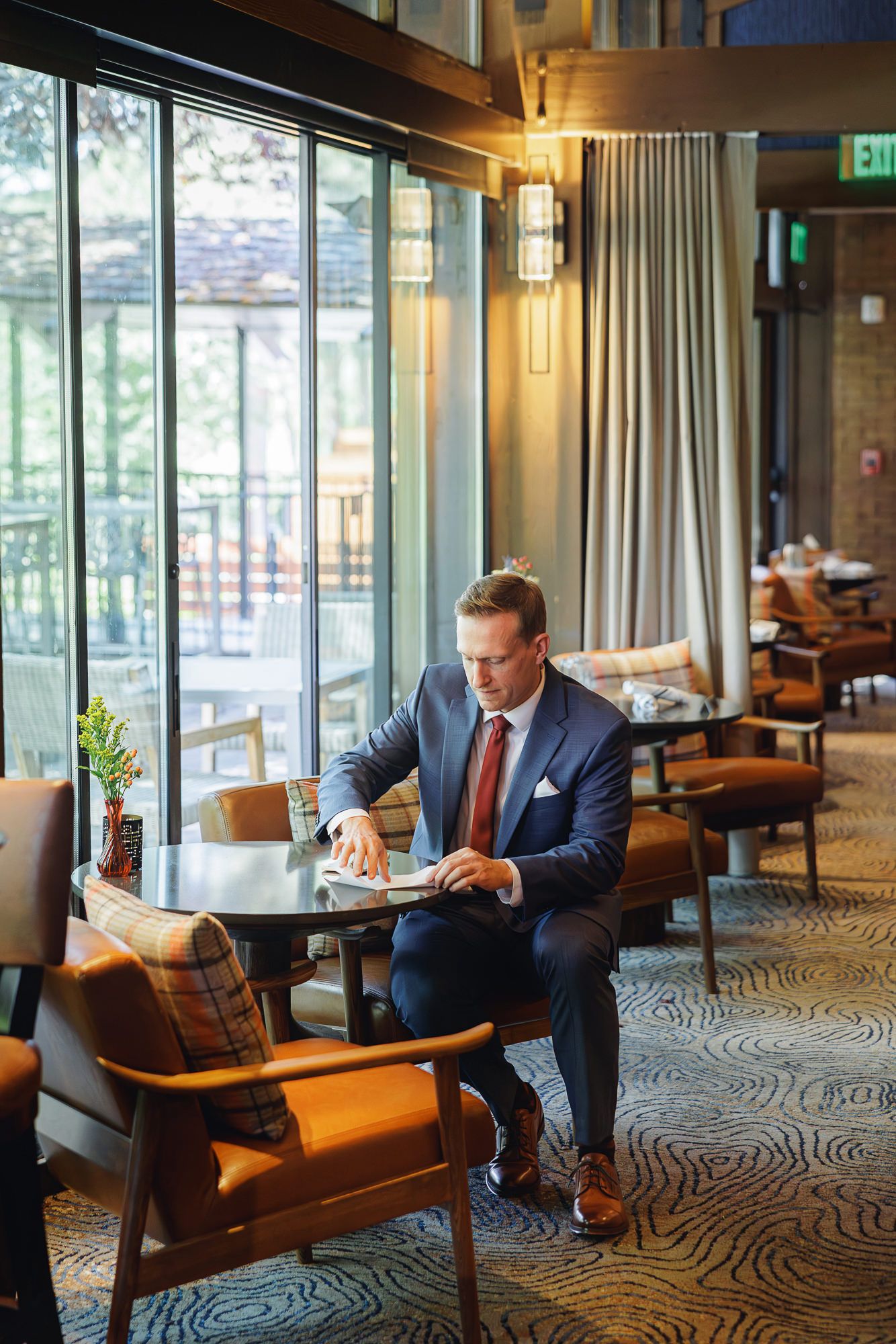 A businessman in a suit sits alone in an elegant hotel lobby with leather chairs and large windows.
