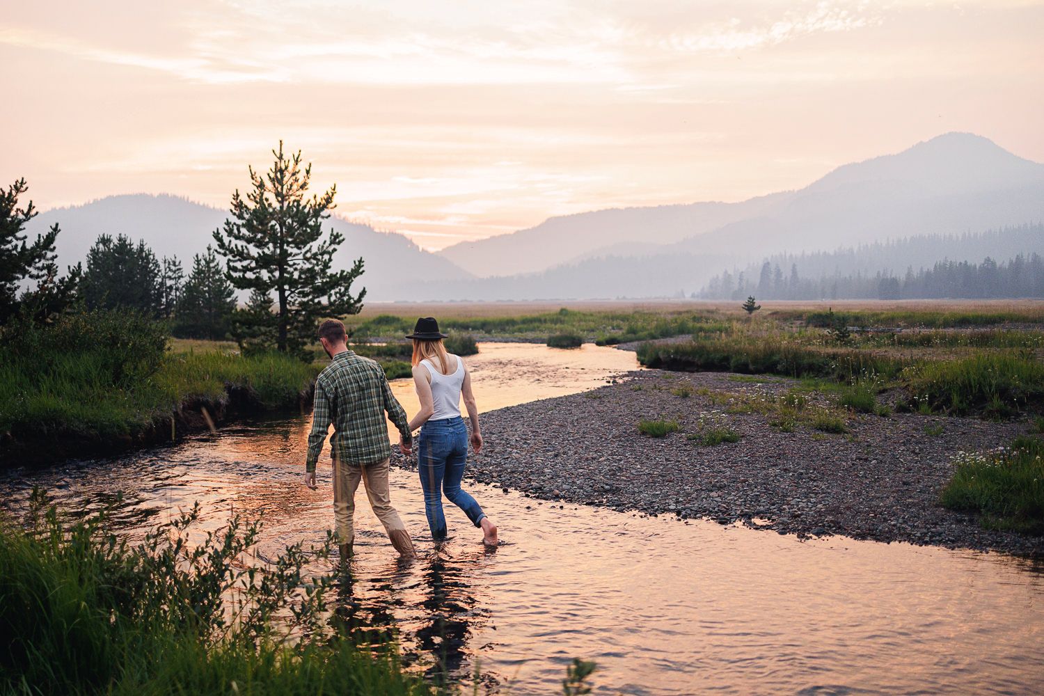 Two people stand in a shallow mountain stream at sunset casting long shadows on the glimmering water.