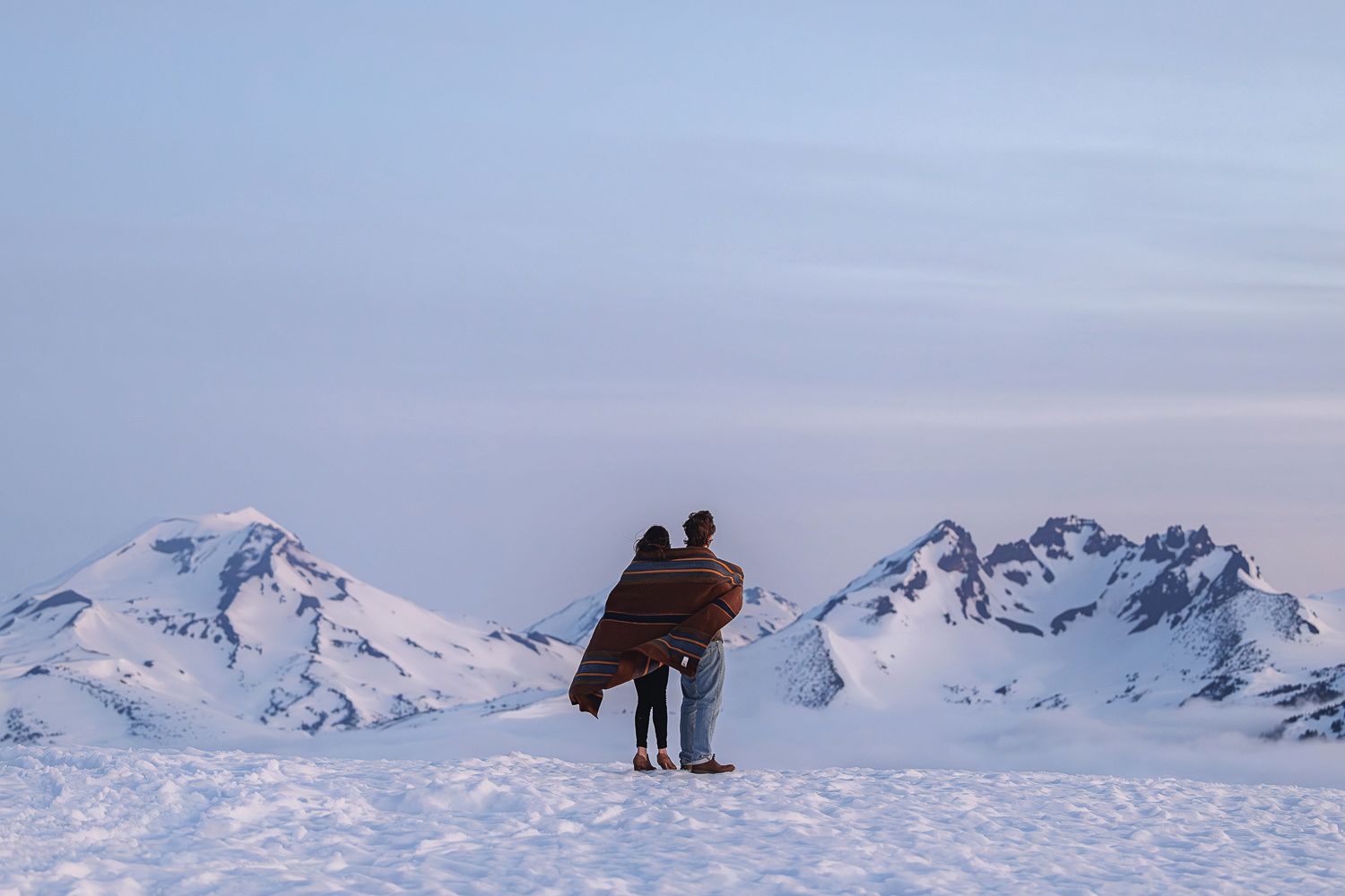 Two silhouettes against a snowy mountain backdrop share an intimate moment above the clouds at dusk.