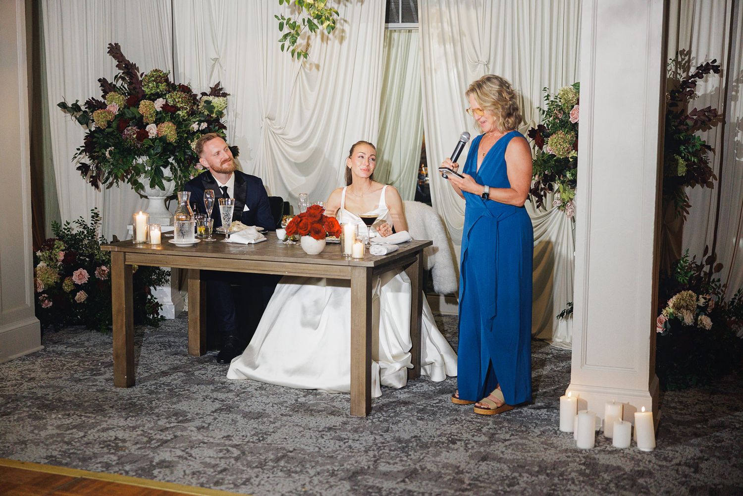 Bride and groom seated at sweetheart table while woman in blue gown delivers wedding toast amid floral arrangements.