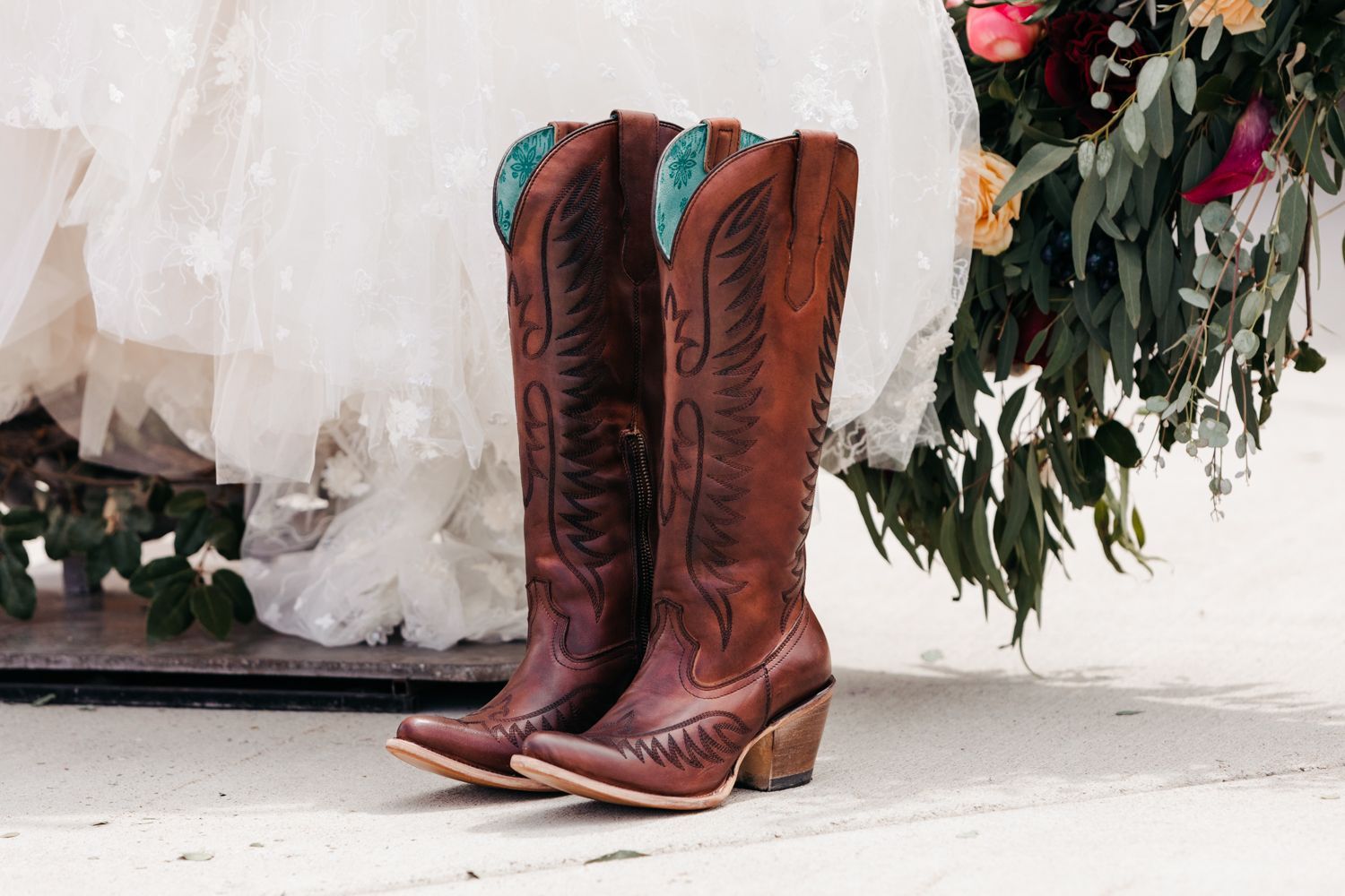 A pair of tall brown leather cowboy boots with decorative stitching sits on a white floor against a white wall.