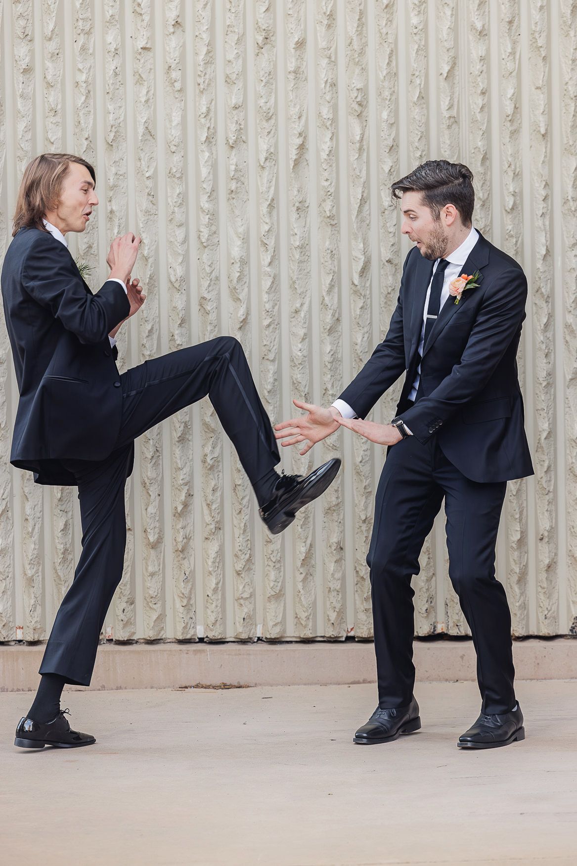 Two people in dark suits playfully interact with one engaging in a high kick move against a textured wall.