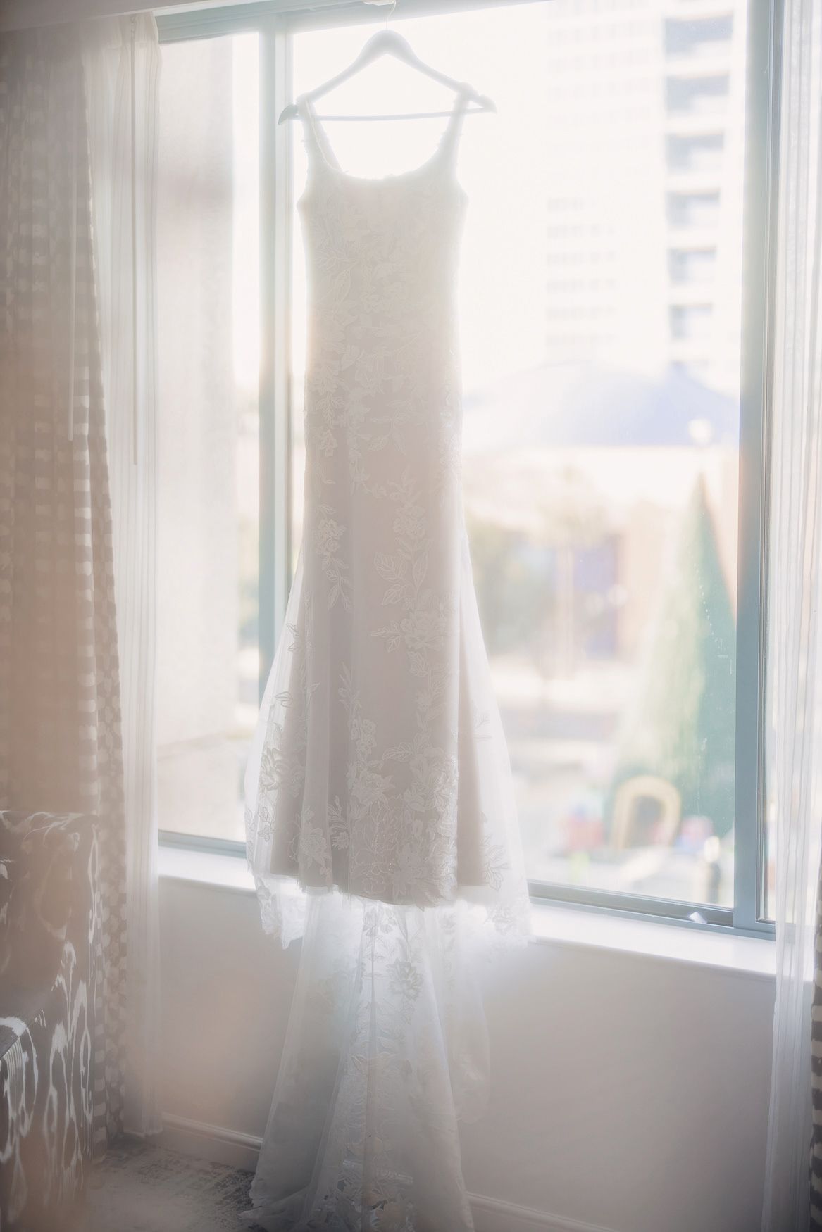A white wedding dress with a long train hangs elegantly in front of a sunlit window creating a dreamy silhouette.
