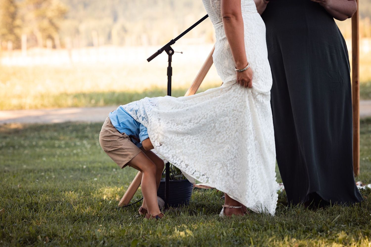 A playful moment during an outdoor wedding ceremony shows a small child peeking under a white lace dress.