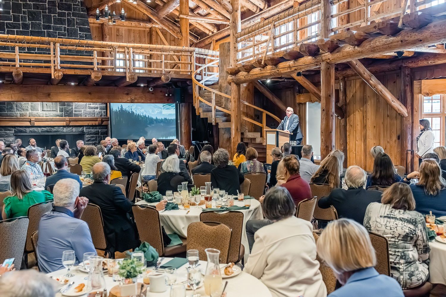Audience seated at round tables in a rustic wooden event venue during a presentation or gathering.