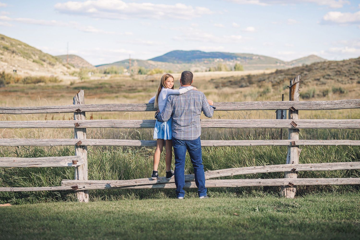 A couple stands at a rustic wooden fence overlooking rolling hills and mountains in the countryside.