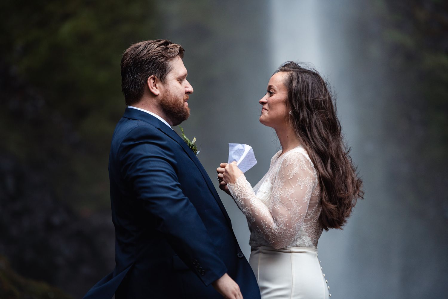 A couple shares a romantic moment in front of a misty waterfall, the bride in white lace and groom in navy suit.