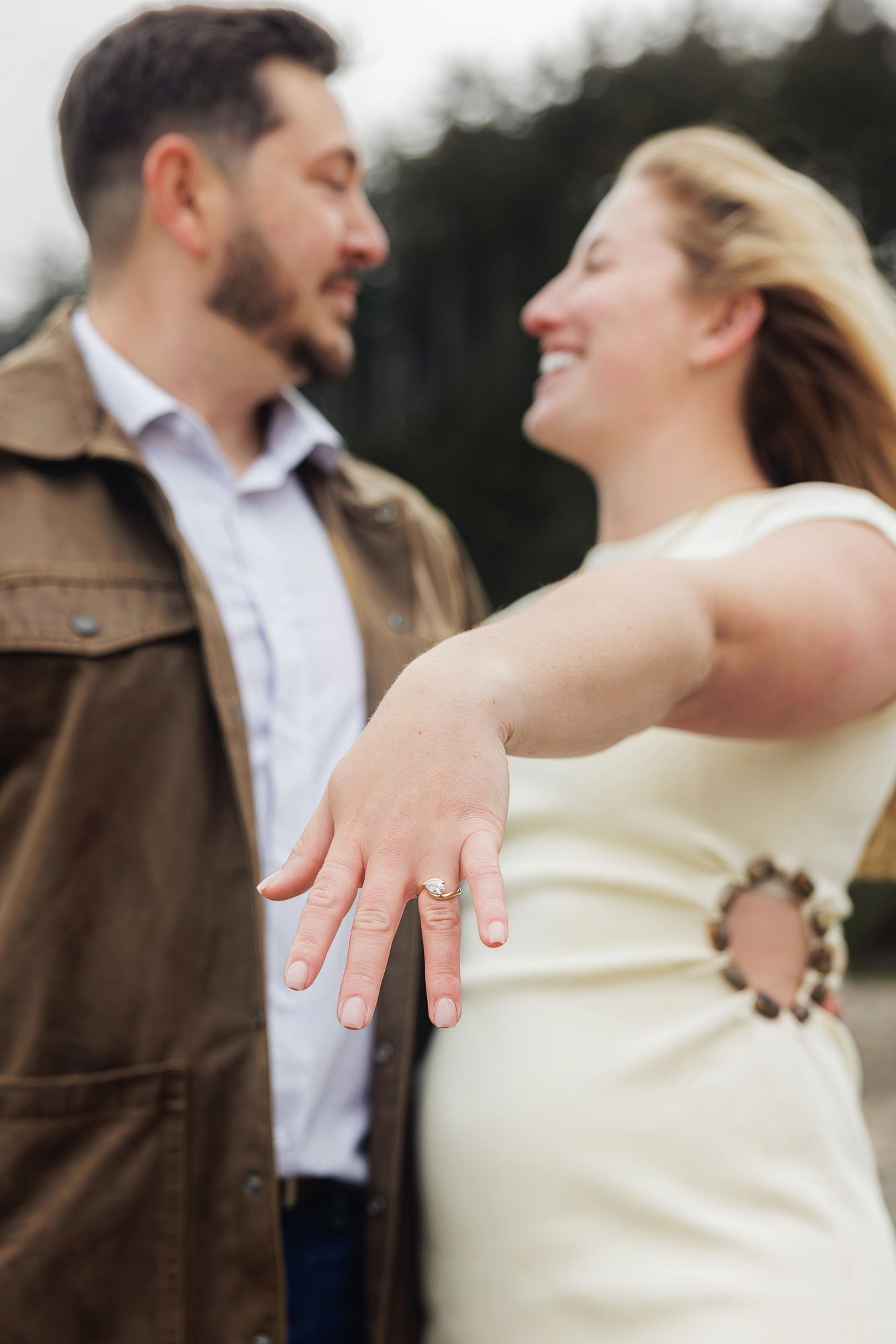 Smiling couple showing off engagement ring, woman in white dress extending hand toward camera, man in brown jacket.