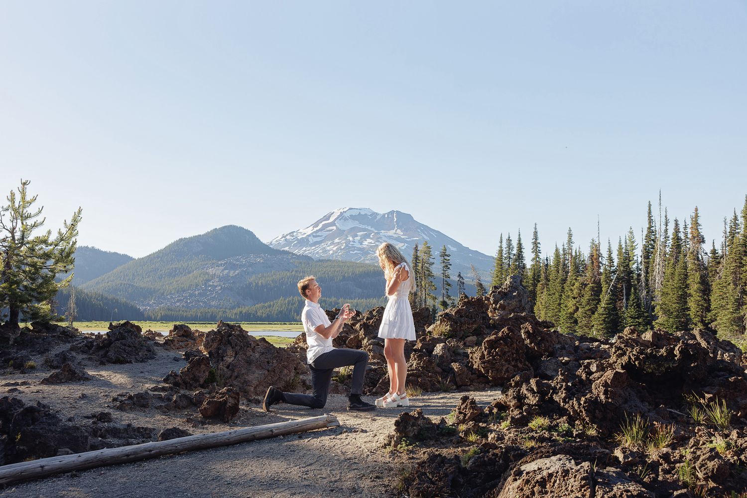 Man proposes to woman on one knee amid volcanic rocks with a snow-capped mountain backdrop in Oregon.