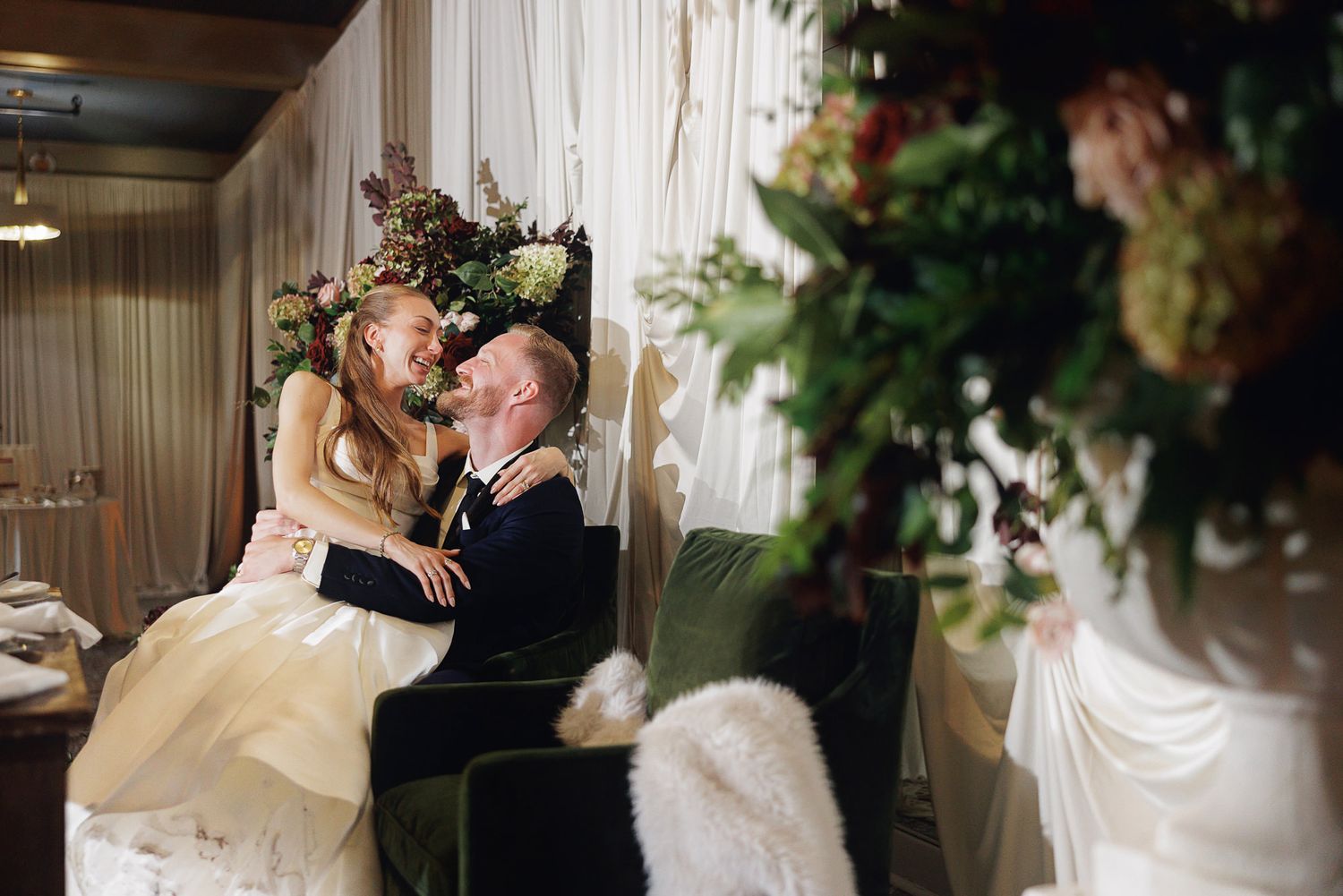 Bride and groom share a joyful moment seated together, surrounded by lush floral arrangements at their wedding reception.