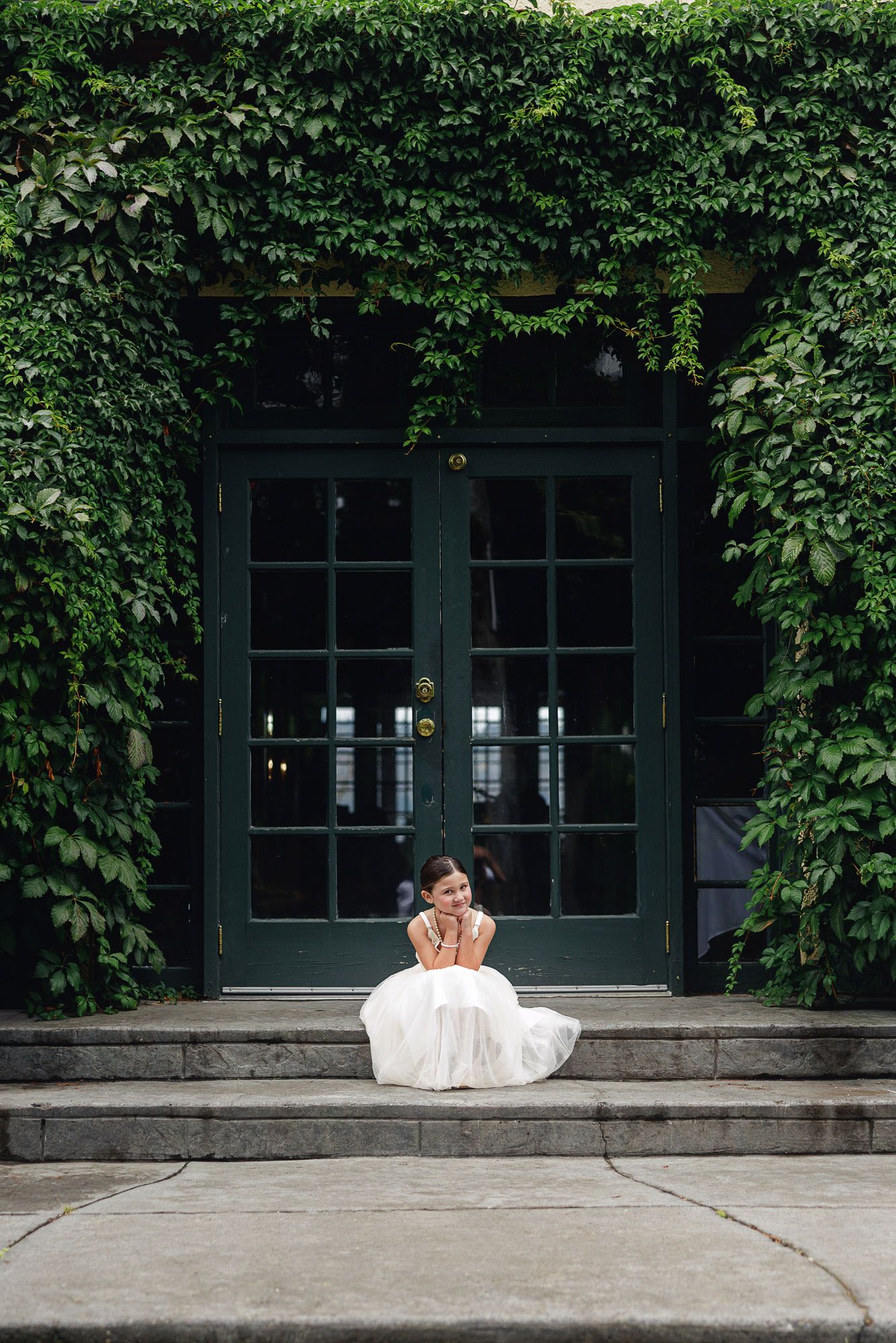 Bride in white dress sitting on stone steps before ivy-covered green French doors.