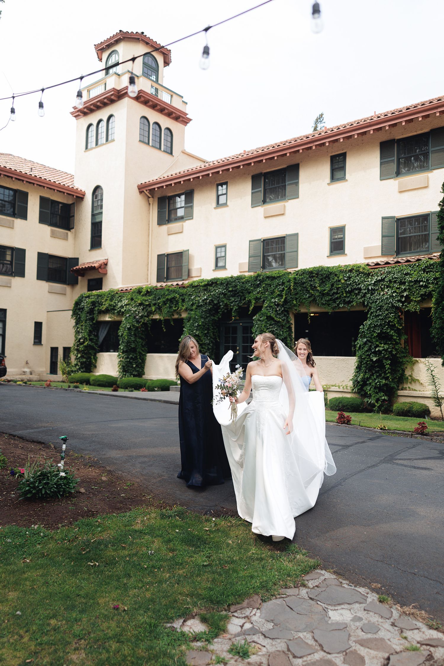 Bride in white gown walks with bridesmaids in black dresses outside an ivy-covered venue on her wedding day.