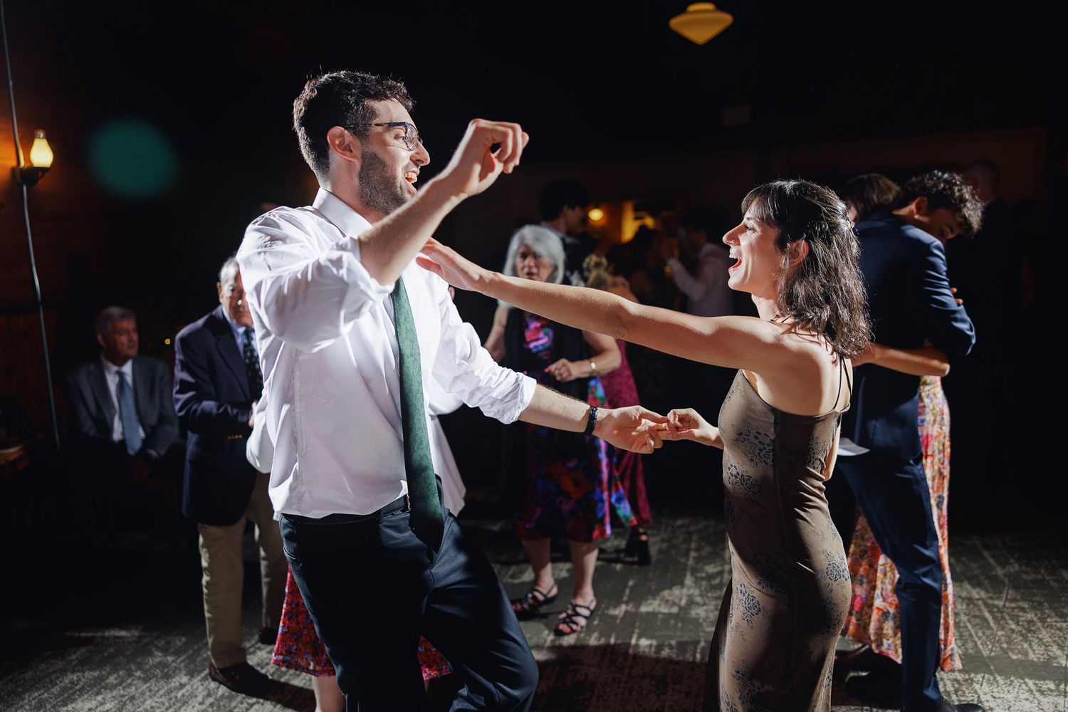 Couple dancing energetically at a wedding reception, man in white shirt and green tie, woman in elegant dress.
