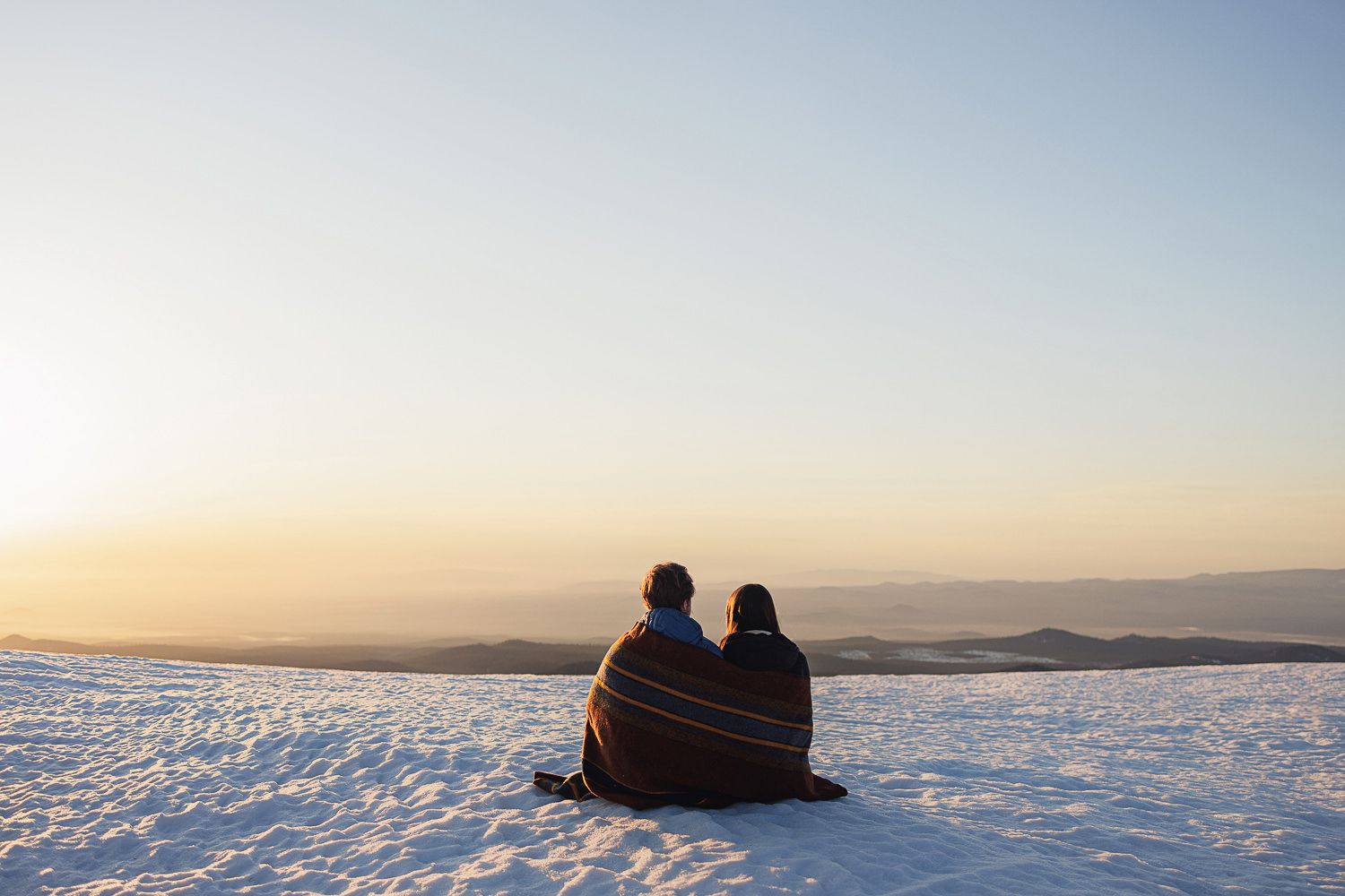 Person sleeps peacefully on snowy ground against vast white winter landscape under pale blue sky at sunset.