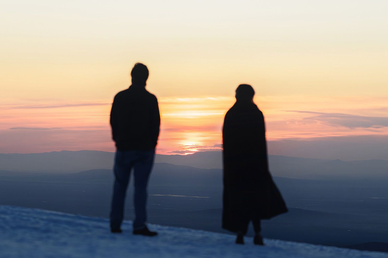 Silhouetted figures stand on a hilltop against a vibrant orange and pink sunset sky.