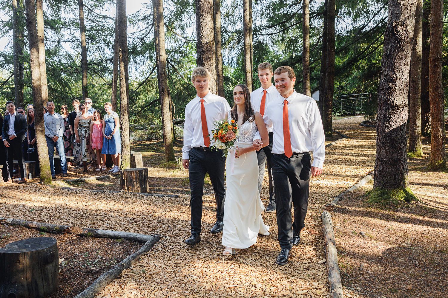Wedding party walks together through a sunlit forest path during an outdoor ceremony.
