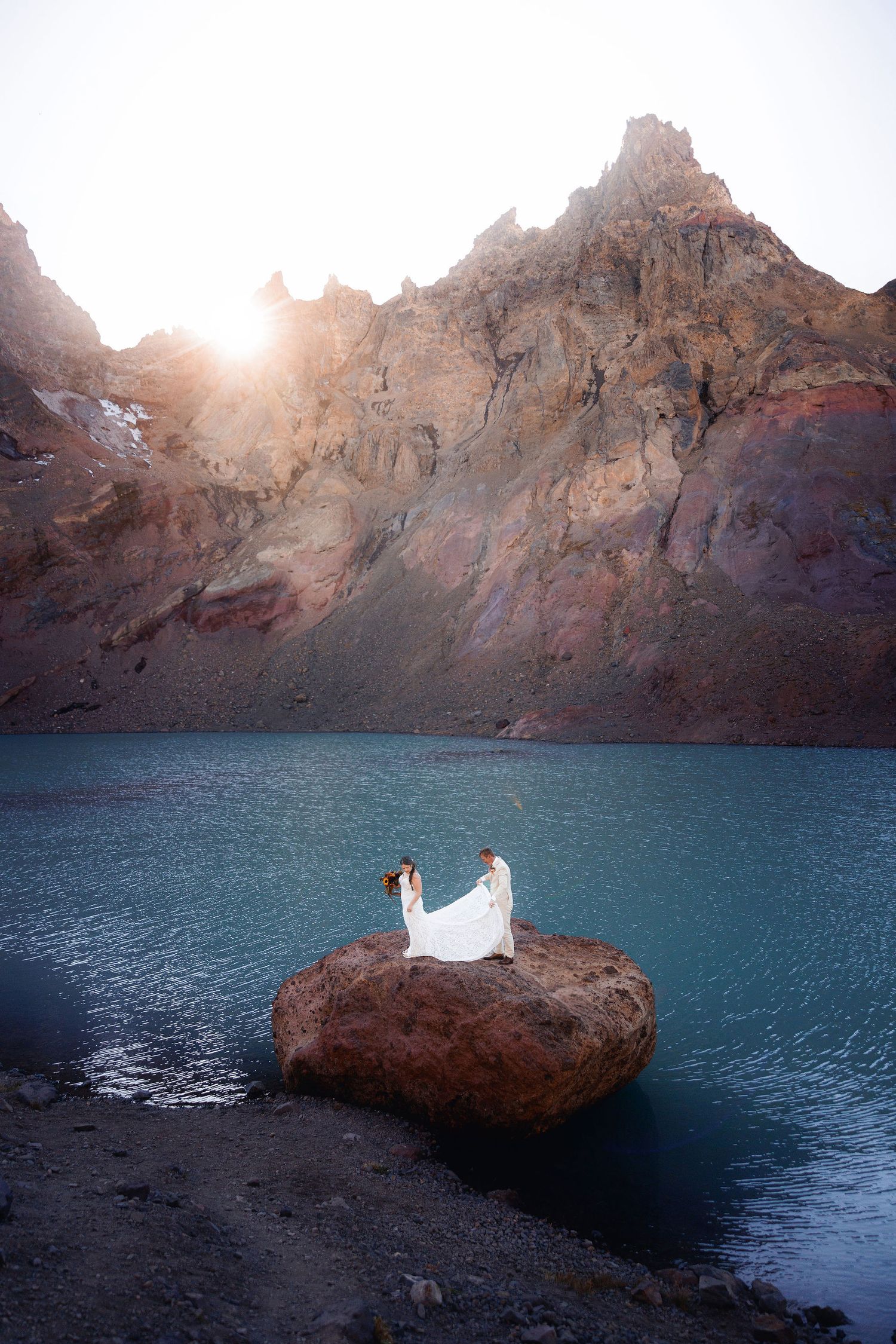 Bride in a flowing white gown and groom stand on a large boulder in a turquoise mountain lake beneath jagged peaks at sunrise.