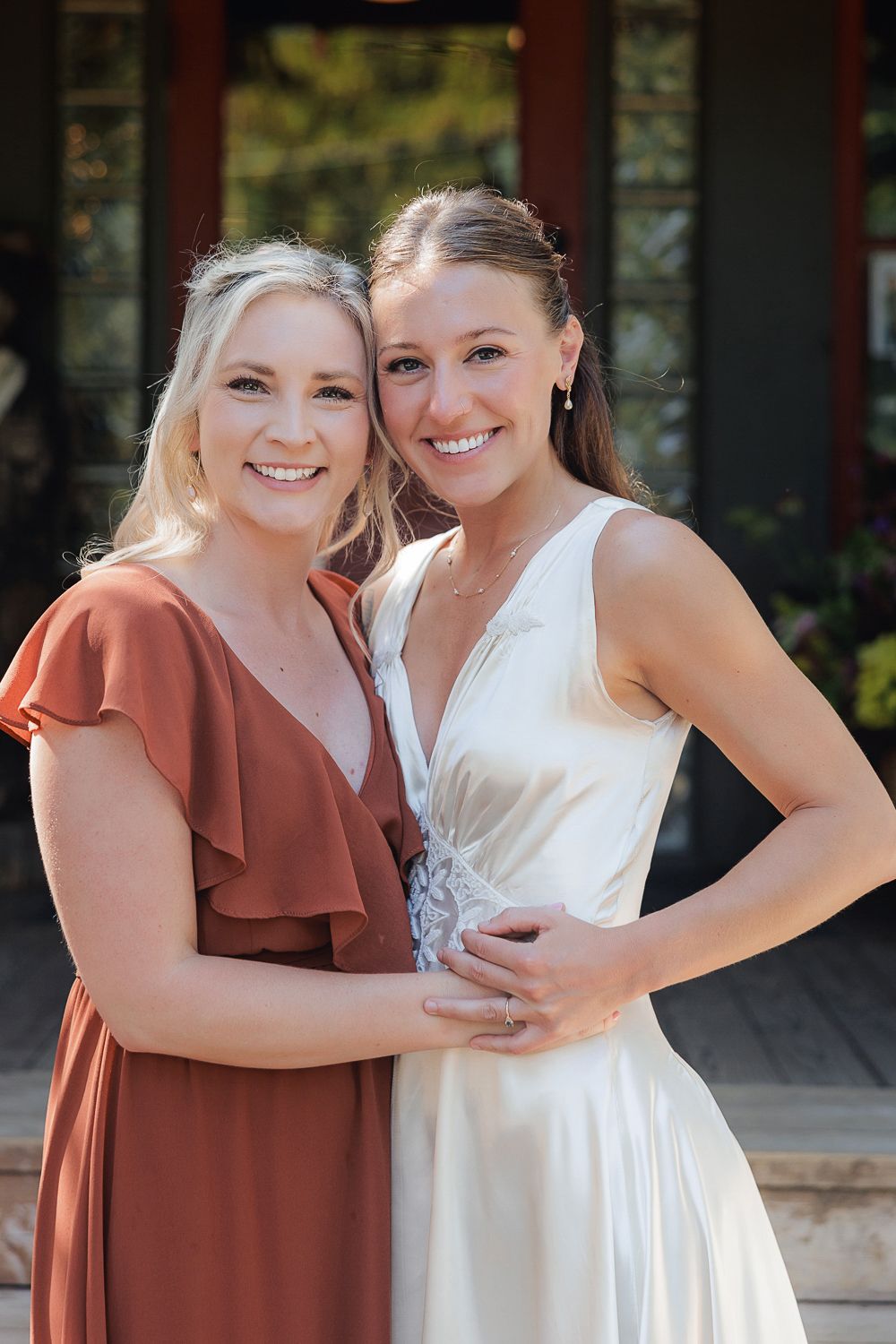 Two friends share a joyful moment in a rustic setting, one wearing a rust-colored dress and one in white.