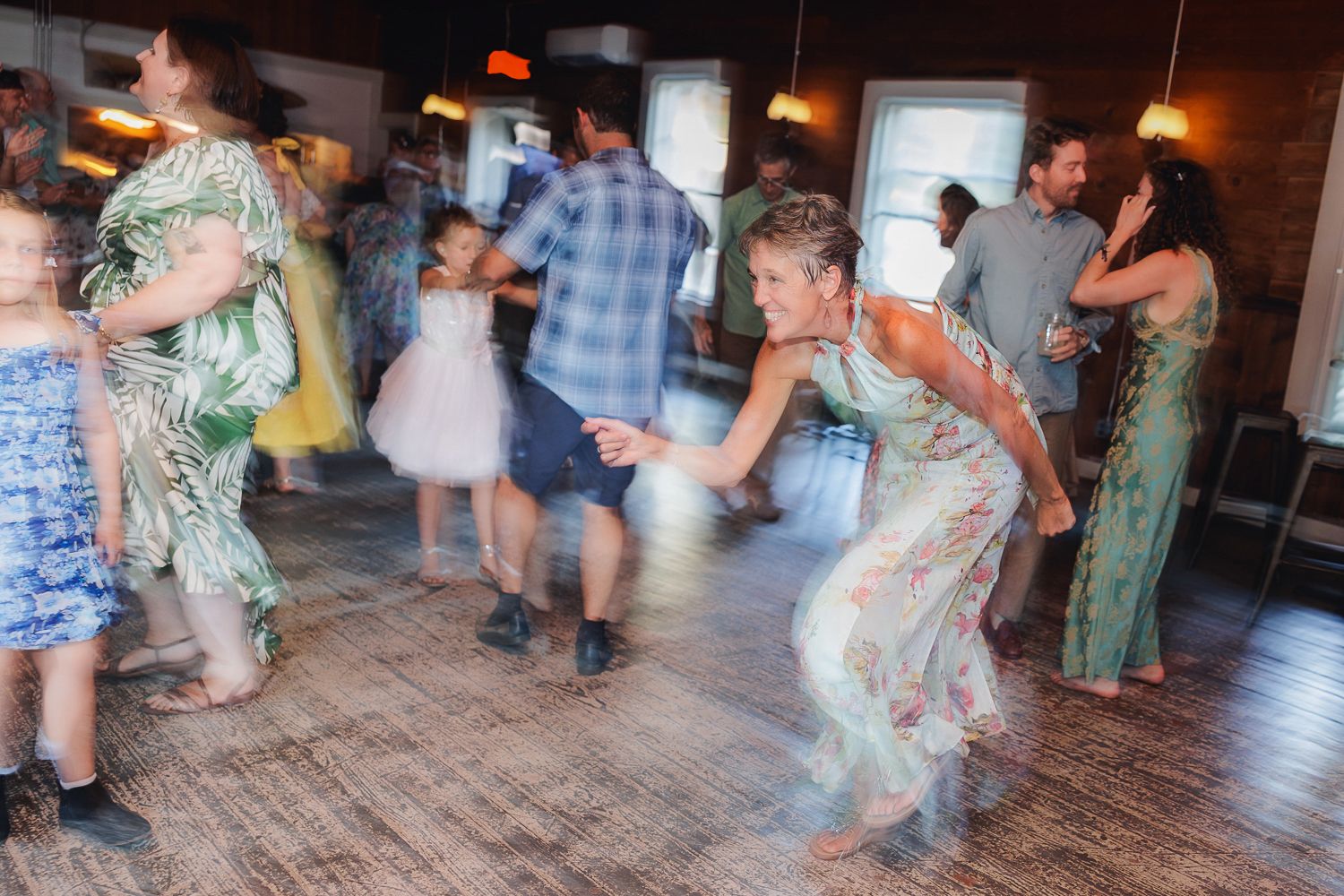 Wedding guests enjoying a lively dance party on a rustic wooden floor illuminated by indoor lighting.