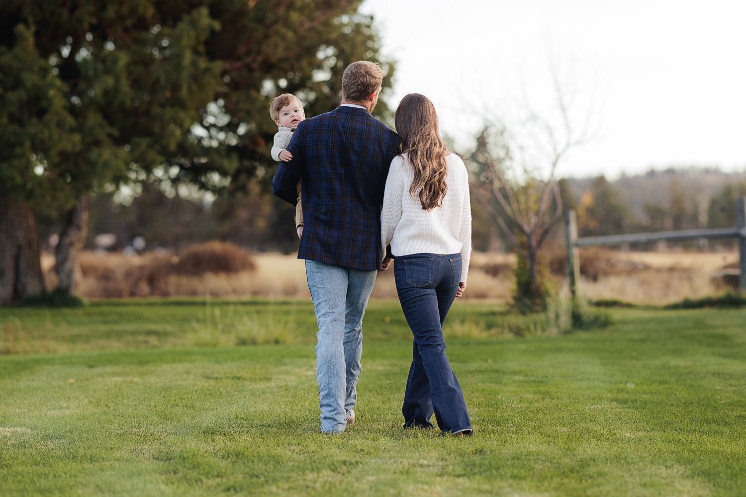 A couple walks together on a grassy lawn while holding their young child on an autumn afternoon.
