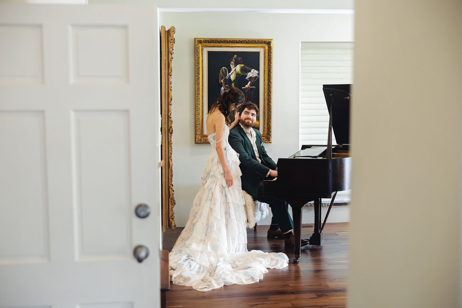 Bride in lace gown stands by groom playing grand piano in elegant room with gold-framed painting.