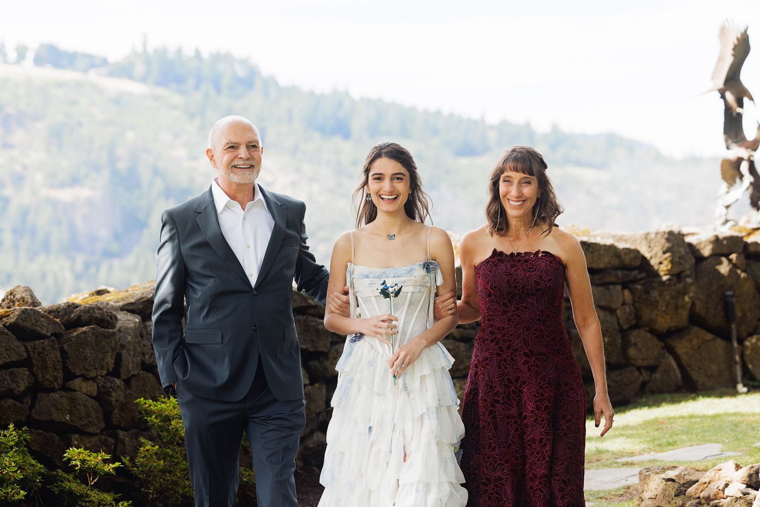 Bride in white gown walks with parents outdoors near stone wall with mountain views.