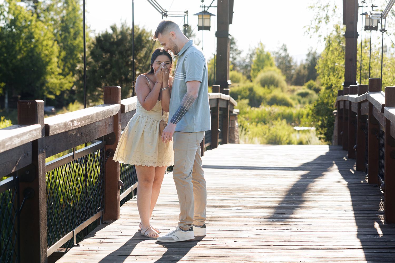 Couple sharing a tender moment on a wooden bridge surrounded by lush greenery in warm sunlight.