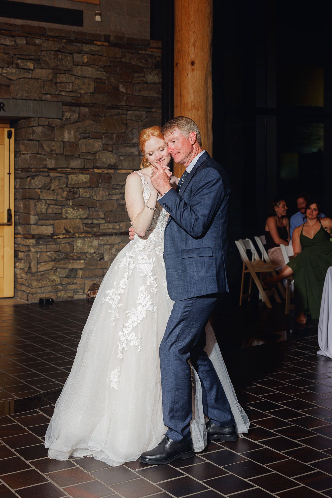 Bride and groom share an emotional first dance in a rustic stone venue, bride in lace gown, groom in blue suit.