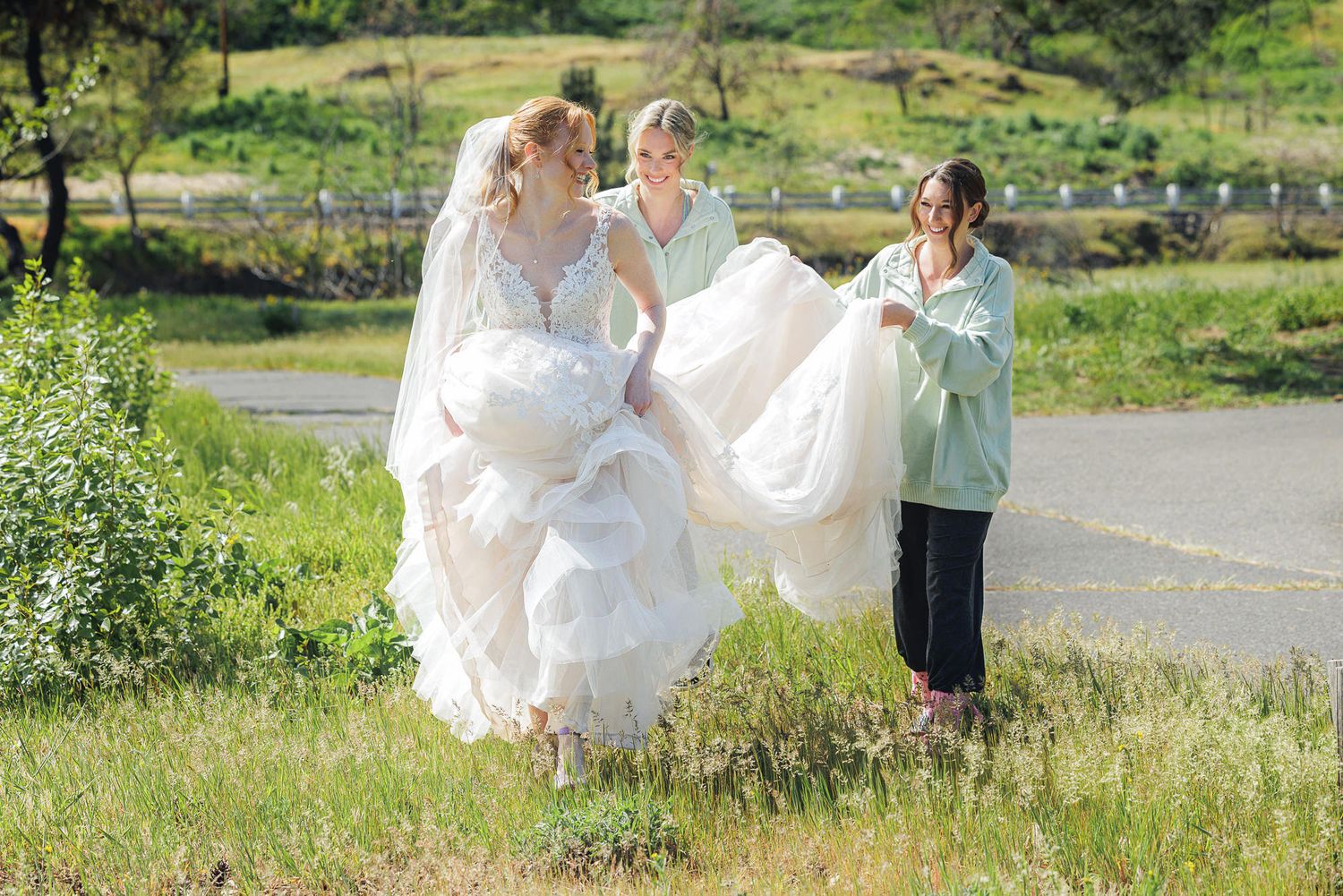 Bride in white gown assisted by two bridesmaids carrying her dress train outdoors on a grassy hillside.