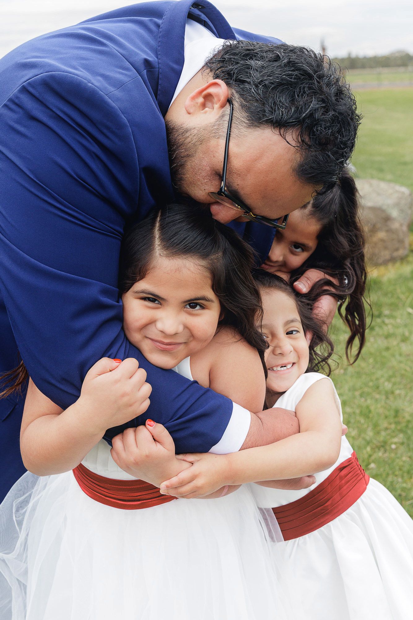 Man in blue suit hugging three young girls in white dresses with red sashes at an outdoor event.