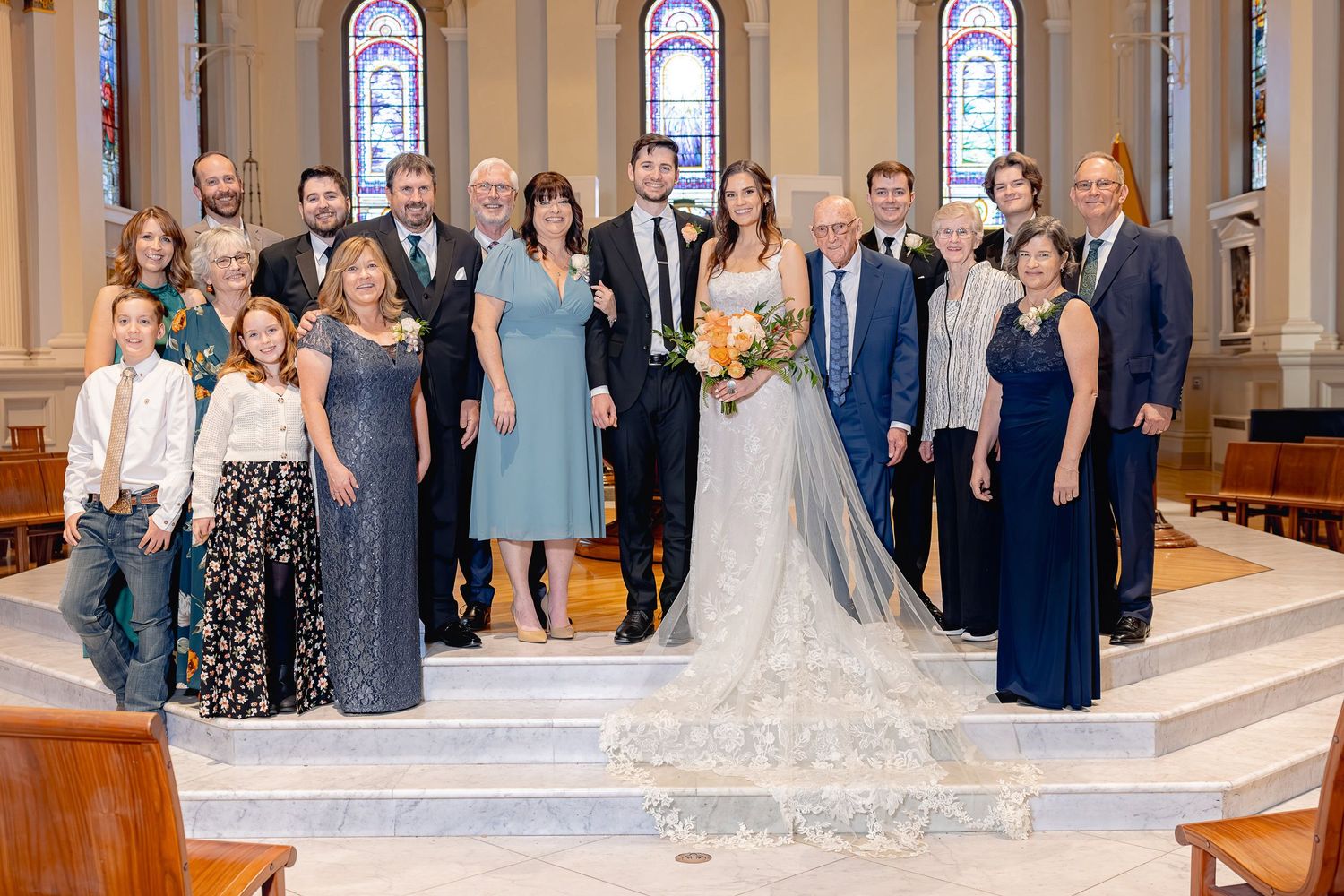 A wedding party in formal attire poses together on the steps of an ornate church with stained glass windows in the background.