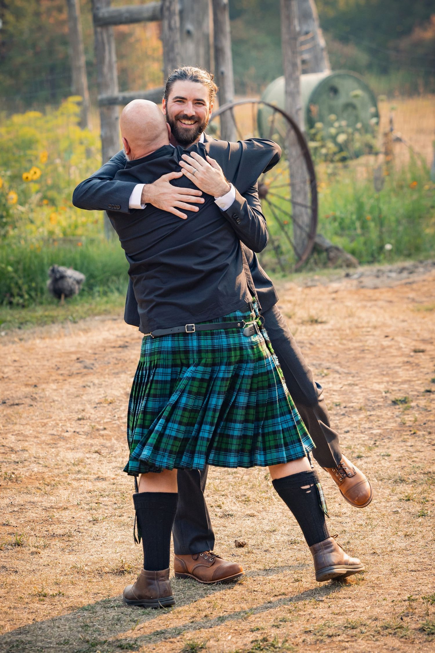 Two people wearing traditional Scottish kilts embrace in a warm hug on a dirt path with rustic scenery in the background.