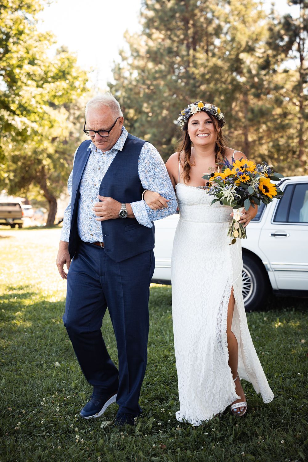 A bride in a lace dress with sunflower bouquet and flower crown walks arm-in-arm with companion in navy suit outdoors.