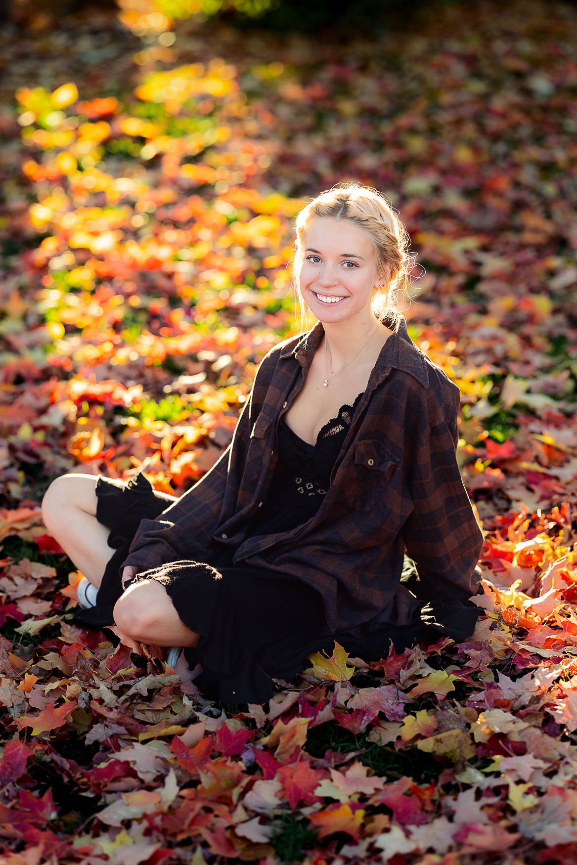 Figure seated among vibrant autumn leaves in black clothing during golden hour.