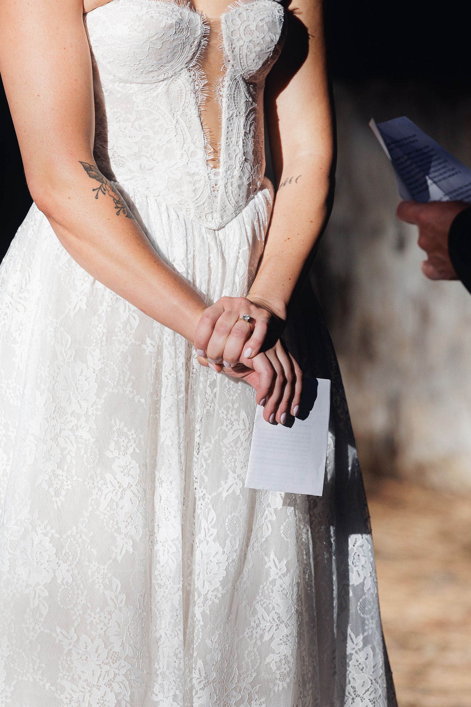 Two brides holding hands during wedding ceremony, wearing white lace gown with tattoos visible, holding vow cards.