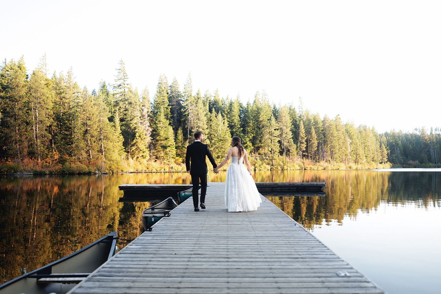 Bride and groom walking hand in hand on a wooden dock overlooking a calm reflective lake surrounded by autumn forest.