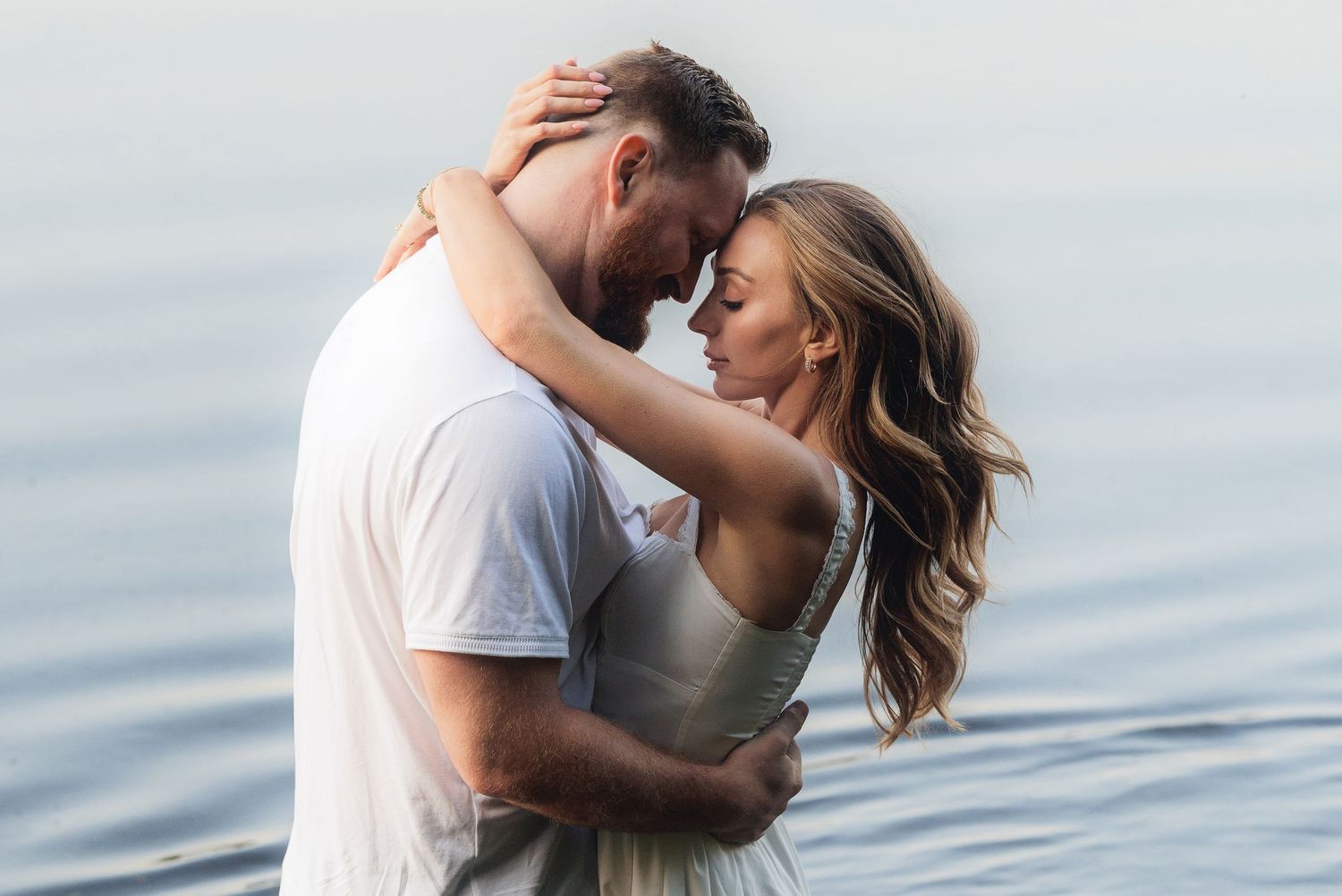 Romantic couple embracing tenderly by calm water, sharing an intimate moment in soft natural light.