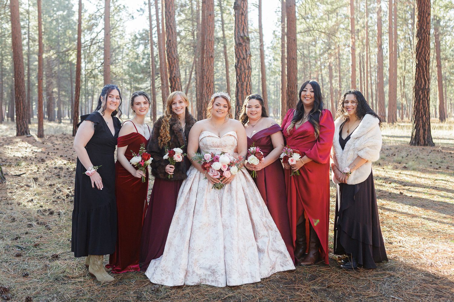 Wedding party in burgundy and black dresses poses together in a sunlit forest of tall pine trees.