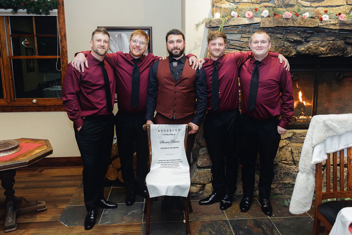 Groomsmen in burgundy attire pose together in front of a stone fireplace at a wedding reception.