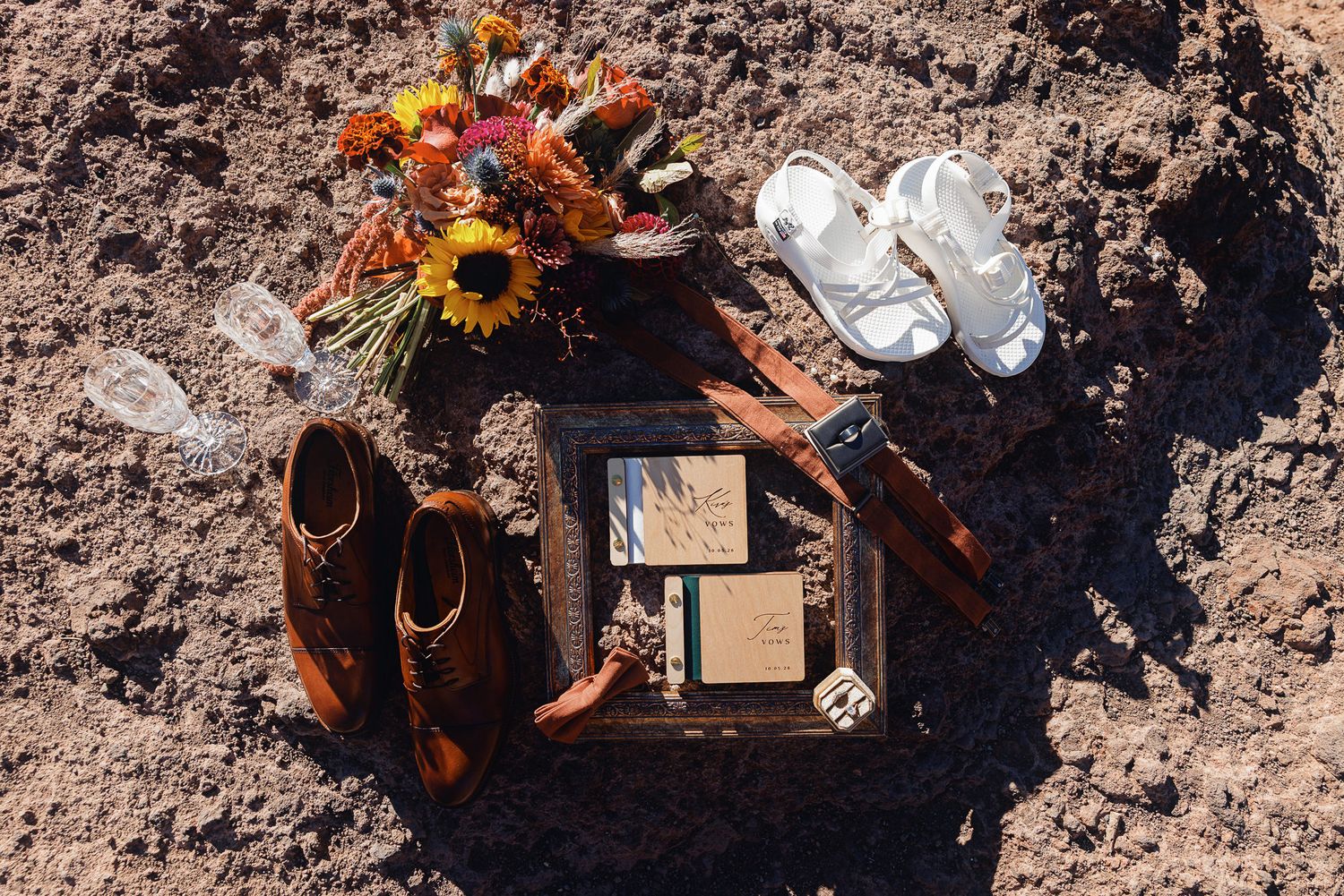 Brown leather men's shoes, white baby sandals, a sunflower bouquet, champagne flutes and framed vows arranged on rustic desert ground.